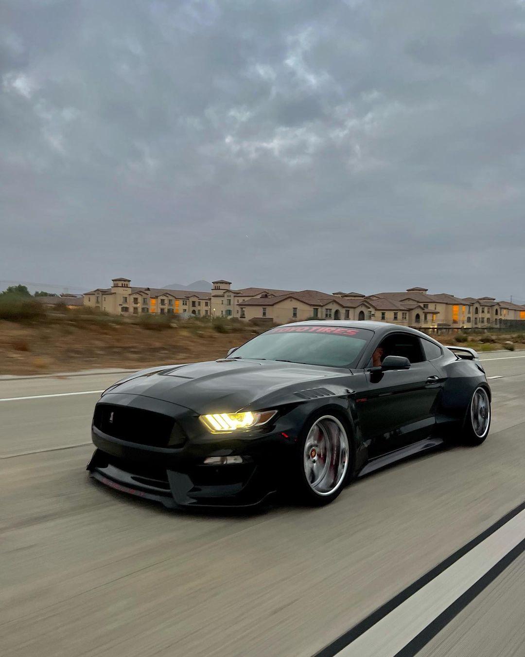 Black Ford Mustang S550 with SHIROKAI widebody kit driving on road under cloudy sky