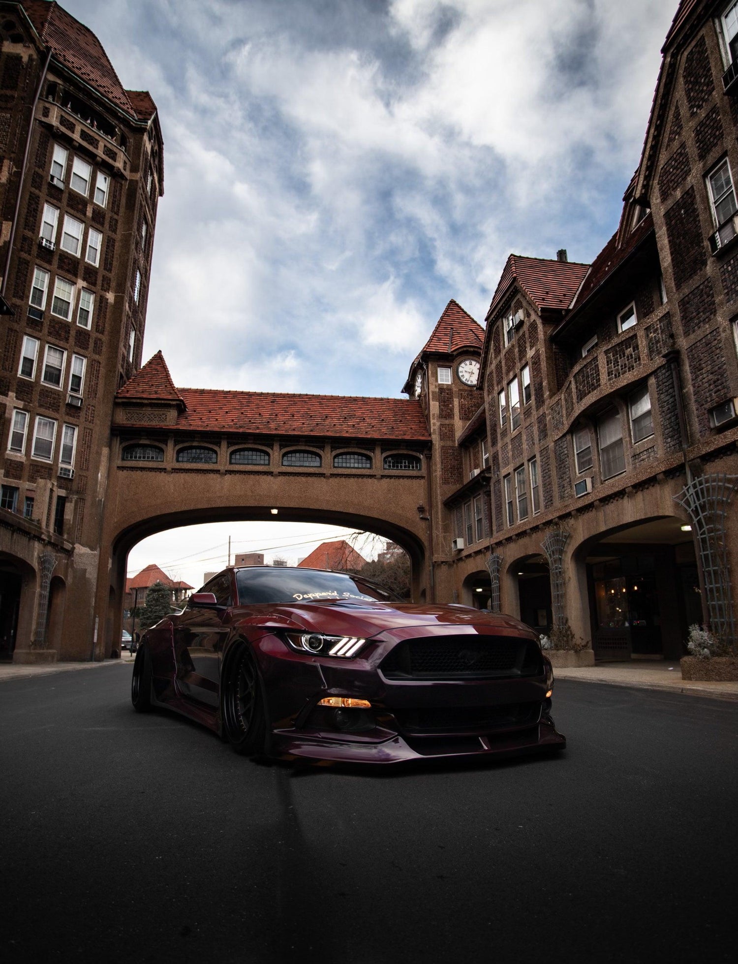 Ford Mustang S550 widebody kit SHIROKAI installed on a dark red Mustang parked in an urban street under a historic arch
