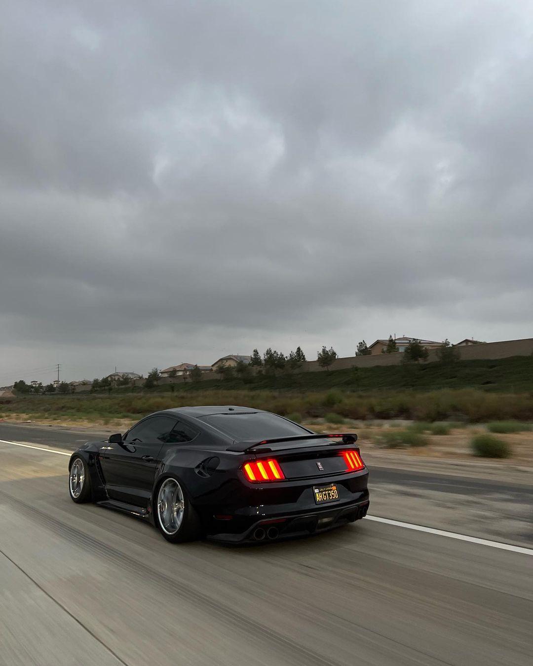 Black Ford Mustang S550 with Ford Mustang S550 widebody kit SHIROKAI driving on highway under cloudy sky
